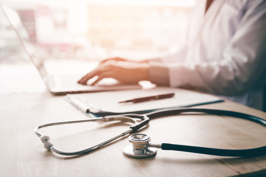 Physician typing on computer next to stethoscope.